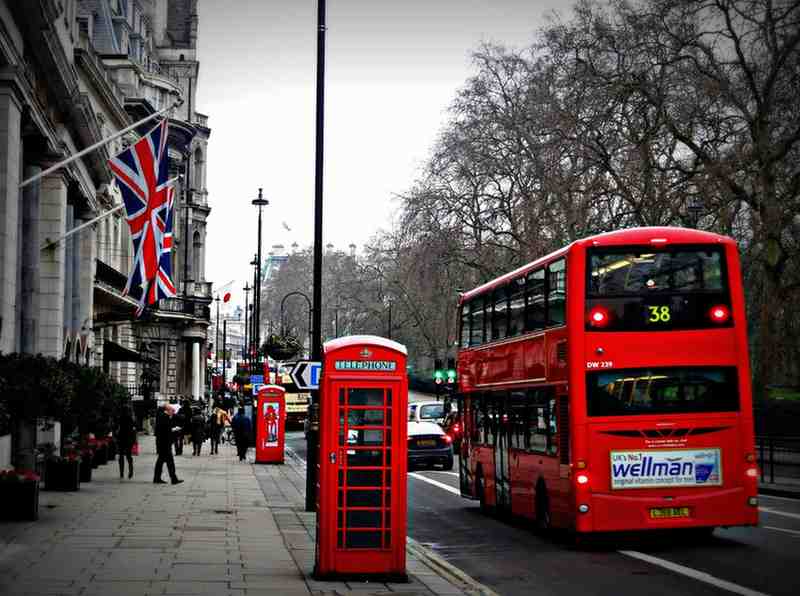 Red double-decker bus and classic telephone booths on a street in London, UK, with Union Jack flag and pedestrians, showcasing iconic British landmarks and city travel scene